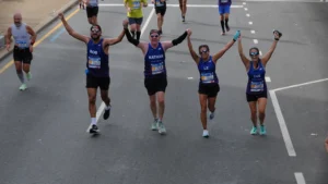 Five runners cross a wide road together with arms linked and raised in celebration.