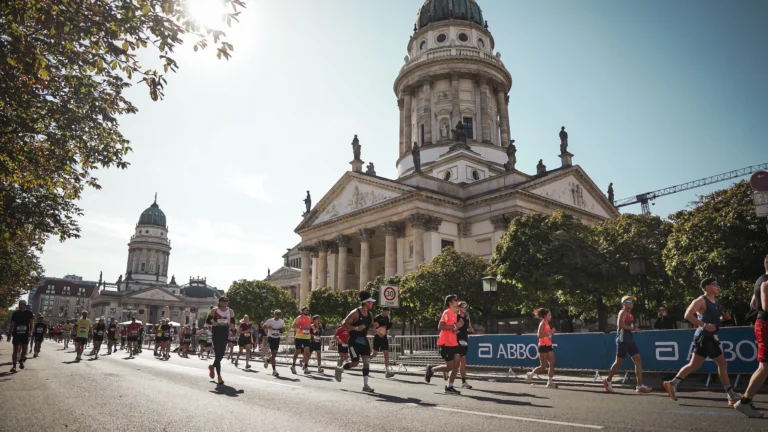 Runners spread across a wide Berlin street past the domed cathedral at Gendarmenmarkt, morning sun coming through the trees, Abbott barriers along the course.